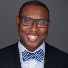 This is a headshot photo of Professor Quayshawn Spencer, a black cis-man. He is wearing black, rectangle glasses and is smiling with his teeth. He is in a dark suit jacket with a light blue shirt and blue bow tie.