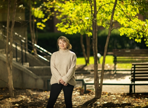 Student Maja Sidzińska is standing slightly left of center of this photo with her hands clasped in front of her waist. She is a white cis-woman with blonde hair tied in a low bun, with bangs. She is wearing a light beige sweater with black pants. The background of the photo is blurred. It contains small trees and a stairway on Penn's campus.